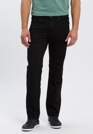 Man die zwarte rechtvallende spijkerbroek, een petrolkleurig T-shirt en zwarte sneakers draagt, staat tegen een effen achtergrond.