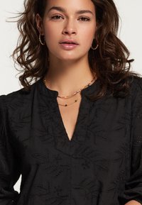 Bluză neagră cu model brodat în frunze, având decolteu în V și mâneci bufante. Purtați cu coliere colorate din mărgele cu mai multe fire.