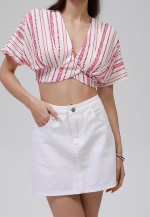 Vrouw die een rood-wit patroon cropped top draagt met een gedraaide voorkant en een witte denim minirok voor een effen achtergrond.