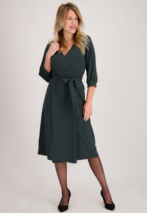 Wikkeljurk van donkergroene stof, met driekwart mouwen, een ceintuur in de taille en een uitlopende, knielange snit. Gladde textuur.