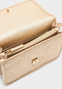 Sac à main texturé en or avec une finition lisse, comportant une poche zippée et un motif de logo en relief. Accent de fermoir carré à l'avant.