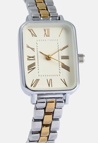 Reloj rectangular con una correa de metal en tonos plateado y dorado. Esfera color crema con números romanos dorados y manecillas. Incluye una corona en el lateral.
