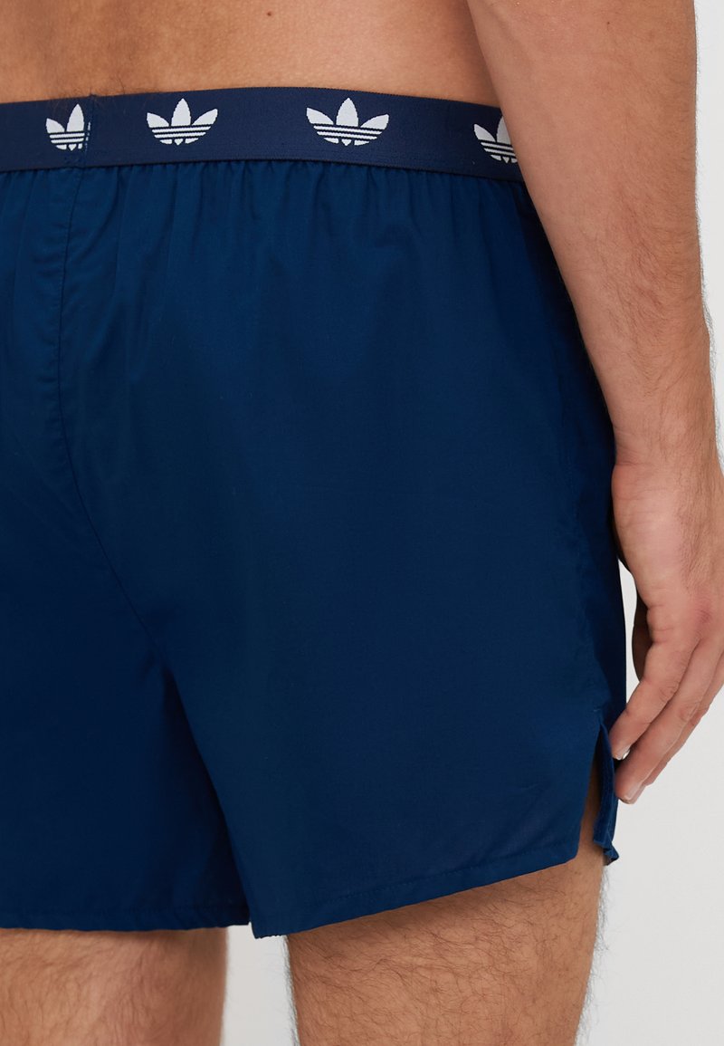 Mørkeblå badebukser med en taljeelastik, der har hvide Adidas trefoil-logoer; inkluderer sideskæringer for bevægelighed og en glat overflade.