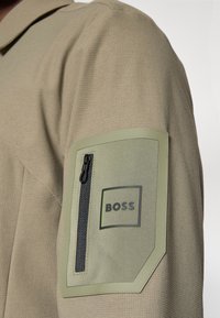 Ανοιχτό χακί μπουφάν με υφή υφάσματος, με έντονη πράσινη τσέπη πατς, μαύρο φερμουάρ και λογότυπο "BOSS" σε τετράγωνο σχέδιο.