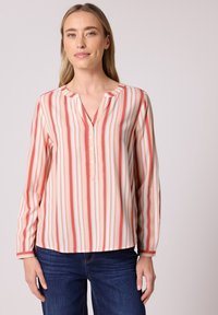 Femme aux cheveux blonds portant un chemisier à manches longues à rayures verticales roses et blanches et un jean bleu sur un fond uni.