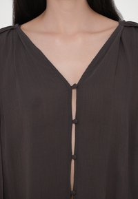 Gros plan d’un chemisier marron foncé texturé avec des boutons recouverts de tissu et un décolleté en V montrant le haut de la poitrine et les clavicules.