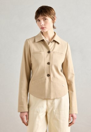 Veste structurée beige avec un col, quatre poches et fermeture à boutons. Le tissu lisse et la coupe ajustée renforcent son design moderne.