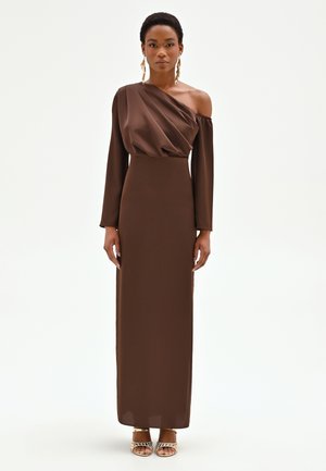 Femme portant une robe asymétrique marron foncé à manches longues, des boucles d'oreilles dorées et des talons ouverts, debout devant un fond blanc.