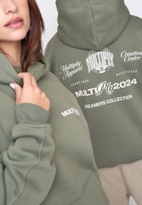 Olivovo zelená mikina s voľným strihom, obsahujúca biele potlače a text, vrátane "Multiply Apparel" a "Dreamers Collection."