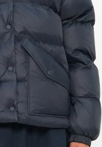 Mornarska puffer jakna z teksturirano površino, ki ima gumbasto zapiranje na velikem, nagnjenem žepu in elastične manšete.