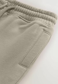 Beige sweats, der har en blød tekstur, elastisk talje med snøre og en sidelomme. Stoffet har et subtilt væve mønster.
