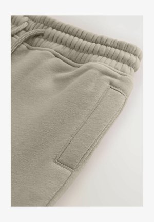 Beige sporta bikses ar mīkstu tekstūru, elastīgu jostu ar auklu un sānu kabatu. Audumam ir smalka auduma raksts.