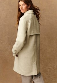 Vrouw met lang bruin haar, gekleed in een beige visgraat wollen jas en grijze broek, staand tegen een effen bruine achtergrond.