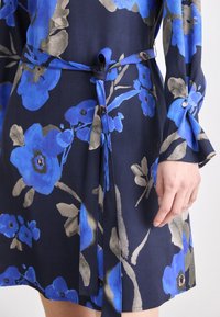 Blommig klänning med en marinblå bakgrund, stora blå blommor och gr å detaljer, knuten i midjan med ett matchande bälte. Långa ärmar med knappdetalj.