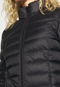 Svart vadderad jacka med hög krage och hel dragkedja. Har quiltade sektioner och slät, blank tygstruktur.