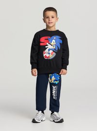 Felpa nera con grafica di Sonic, abbinata a pantaloni della tuta blu navy con design blu di Sonic e testo bianco "SONIC". Scarpe da ginnastica grigie.