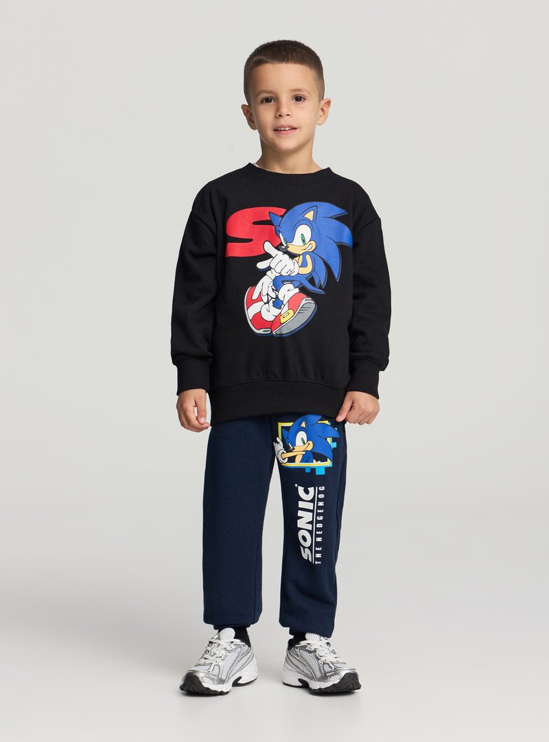 Felpa nera con grafica di Sonic, abbinata a pantaloni della tuta blu navy con design blu di Sonic e testo bianco "SONIC". Scarpe da ginnastica grigie.