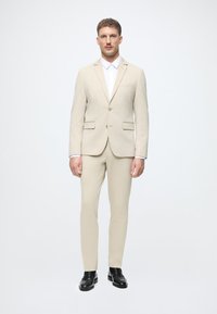 Bežinis švarkas su vienu mygtuku ir siaurais džinsais. Po juo – balta oficiali marškiniai. Juodi loaferiai užbaigia įvaizdį.