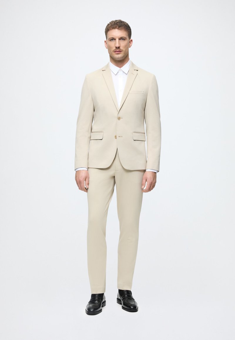 Bežinis švarkas su vienu mygtuku ir siaurais džinsais. Po juo – balta oficiali marškiniai. Juodi loaferiai užbaigia įvaizdį.