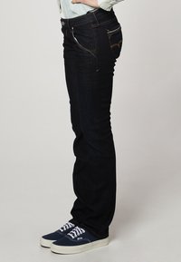 Jeans de mezclilla azul oscuro con corte recto, presentan una cintura de altura media, costuras de color cobre y bolsillos delanteros y traseros. Combinados con zapatillas de deporte azul marino.