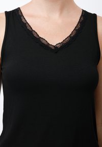 Schwarzes Tanktop mit V-Ausschnitt und gewelltem Spitzenbesatz. Hergestellt aus einem glatten, dehnbaren Stoff. Ärmellos.