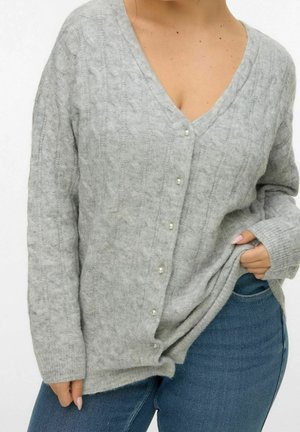 Femme portant un cardigan gris clair en maille torsadée avec des boutons en perle, associé à un jean bleu, tenant l'ourlet du pull d'une main.