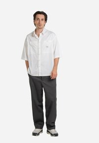 White short-sleeve button-up shirt with two chest pockets, worn with loose-fitting dark gray pants and light-colored sneakers.