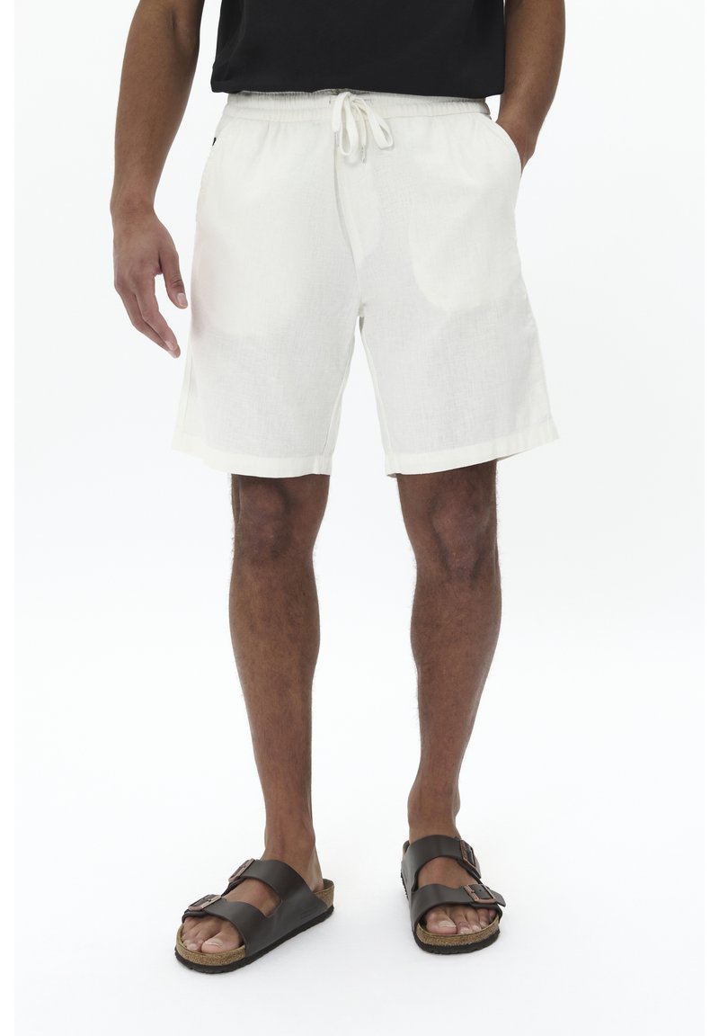 Homme portant un short blanc à cordon, un t-shirt noir et des sandales marron foncé, debout sur un fond blanc uni.