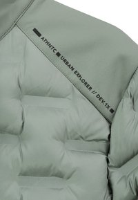 Heller grüner gesteppter Jackenbereich mit schwarzem Text "ATHNTC URBAN EXPLORER // DEV.1X" in der Nähe der genähten Naht.