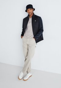 Herremode outfit med en sort dunjakke, beige sweater, creme bukser og hvide sneakers med gummisål. Inklusiv en bucket hat.