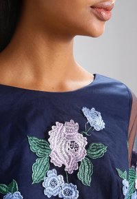 Marinblå klänning i tyg med intrikata blommönster i lila, blå och grön, med en vid halsringning och genomskinliga ärmar.