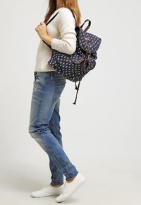 Mochila navy con estampado de ancla blanca, cierre de cordón, detalles en marrón y solapa. Llevada en un hombro, combinada con vaqueros de color azul claro y zapatos blancos.