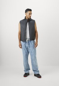 Black, quilted sleeveless vest worn over a white tank top, paired with light blue denim pants and black shoes with red accents.