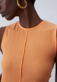 Ribbed, sleeveless dress in peach-orange fabric with subtle vertical lines; features a round neckline and central seam detail. Gold hoop earring visible.