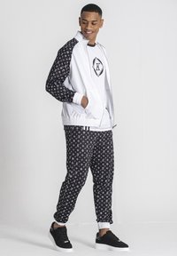 Chándal deportivo con una chaqueta blanca que presenta una manga de patrón negro, combinado con pantalones negros que muestran un patrón de logotipos en repetición. Zapatillas negras.