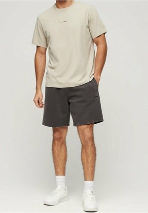 Beige Hemd mit kurzen Ärmeln und minimalem Logo, kombiniert mit dunkelgrauen Shorts. Getragen mit weißen Sneakers und sichtbaren weißen Knö socks.