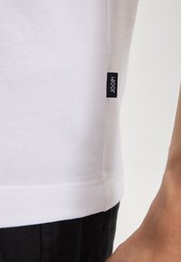 White cotton t-shirt with a rounded hem, featuring a small black tag labeled "JOOP!" on the left side. Smooth texture, simple design.