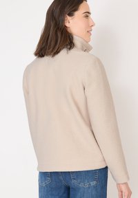 Donna con giacca in pile beige a colletto alto, in piedi di lato, abbinata a jeans blu, su uno sfondo bianco.