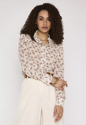 Vrouw met krullend haar draagt een bloemrijke blouse met ruches, crèmekleurige hoge taillebroeken en gouden armbanden en ringen, staand tegen een effen achtergrond.
