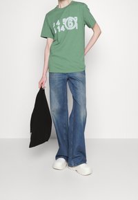 Zelené tričko s bílými potisky čísel, kombinované s modrými rozšířenými džíny a bílými tlustými teniskami; drží černou tašku.