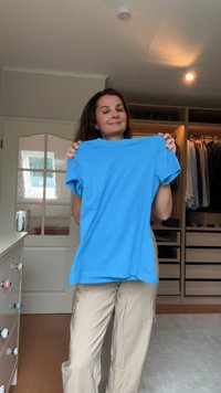 Blue cotton T-paita, jossa on lyhyet hihat ja pyöreä kaula-aukko, pidetään ylhäällä vaaleaa taustaa vasten, ja taustalla näkyy vaatekaappi, joka on täynnä vaatteita.