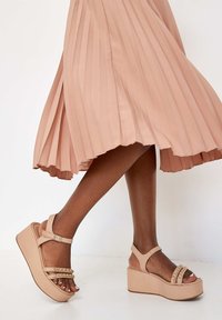 Beige platform sandalen met een chunky zool, voorzien van dunne banden en gouden decoratieve accenten. De banden zijn verstelbaar bij de enkel.