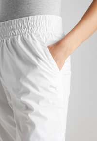 Bílé sportovní kalhoty s elastickým pasem, boční kapsou a nenápadným logem. Hladký materiál, lehká tkanina, vhodné pro aktivní nošení.