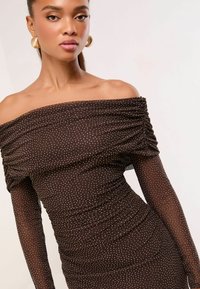 Brūna kleita ar atvērtām plecu daļām un ručiem, piedāvā teksturētu audumu ar zelta punktiņiem. Pieguļošs siluets.