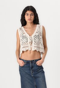 Háčkovaná crop top v off-white s kvetinovými vzormi, výstrihom do V a čipkovým lemovaním; v kombinácii s denimovou sukňou s vysokým pásom.