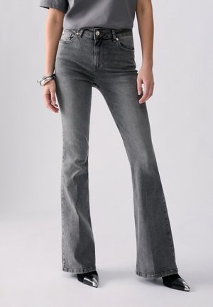 Donna che indossa jeans a vita alta grigi a zampa, scarpe nere a punta e una maglia grigia, in piedi su uno sfondo chiaro uniforme.