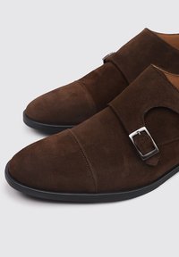 Zapatos de ante marrón con un diseño de puntera puntiaguda, que cuentan con un solo detalle de hebilla metálica y costuras en la parte superior. Suela de goma negra.
