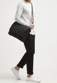 Black messenger bag with flap, minimal design, made of smooth material. Man wearing striped long-sleeve shirt, black trousers, and white sneakers.