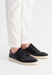 Baskets en cuir noir avec une tige texturée, des lacets plats et des semelles en caoutchouc de couleur claire, associées à un pantalon blanc à coupe ajustée.