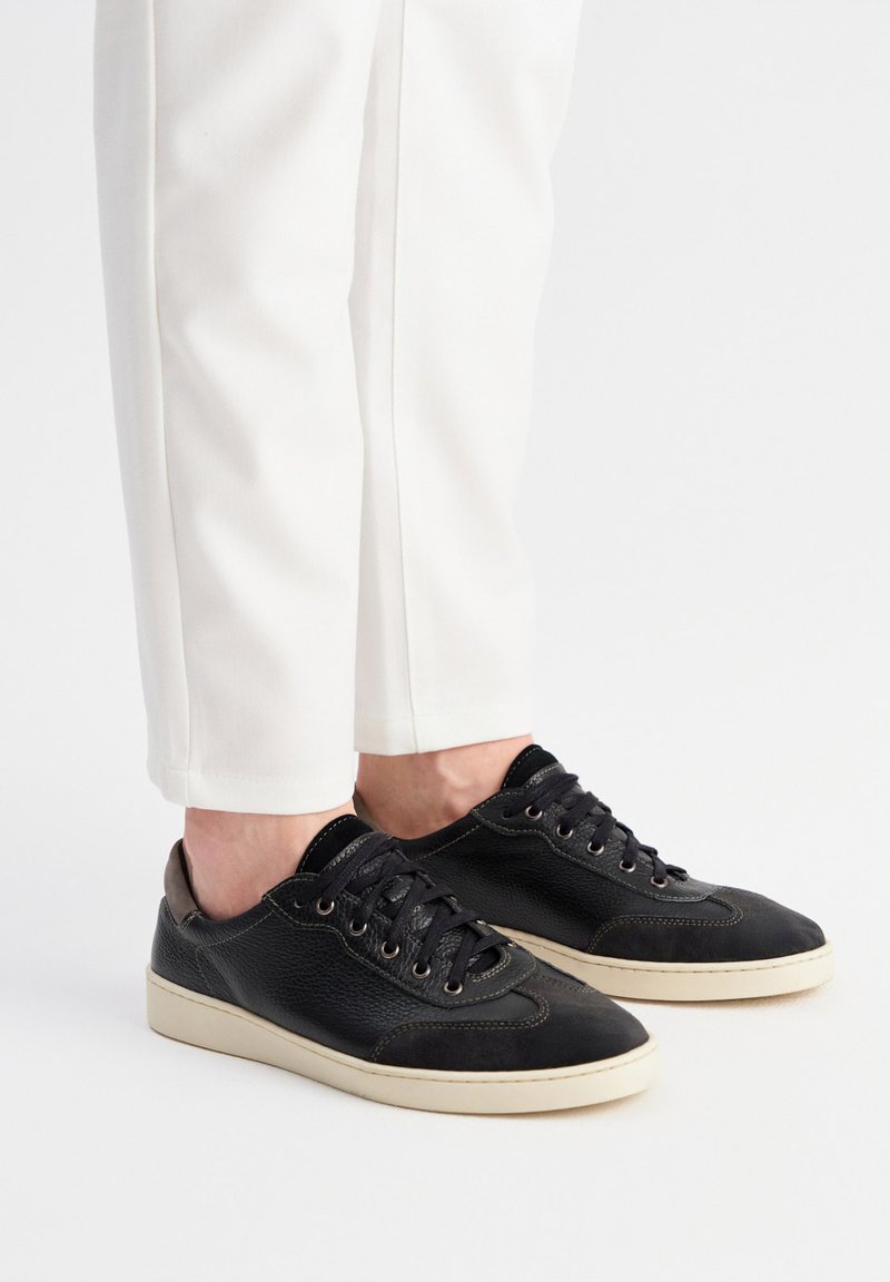Sneakers in pelle nera con tomaia testurizzata, lacci piatti e suole in gomma di colore chiaro, abbinati a pantaloni bianchi a gamba affusolata.